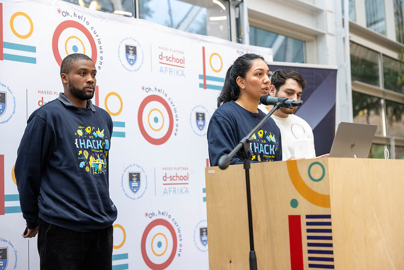 Three hackathon participants stand on stage presenting their project to an audience.