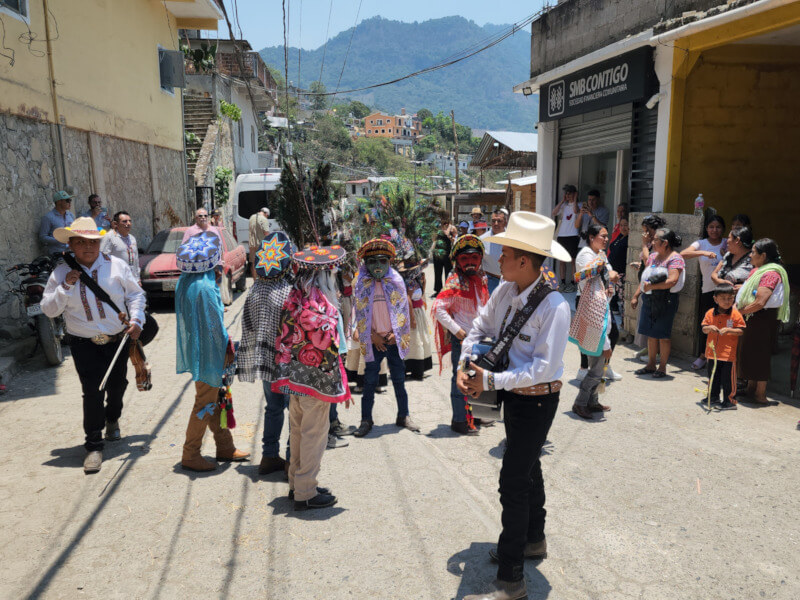 Banner image of a lively street scene in rural Mexico, with people in traditional costumes and masks dancing. A young man stands in the center playing a guitar, surrounded by colorful celebration.